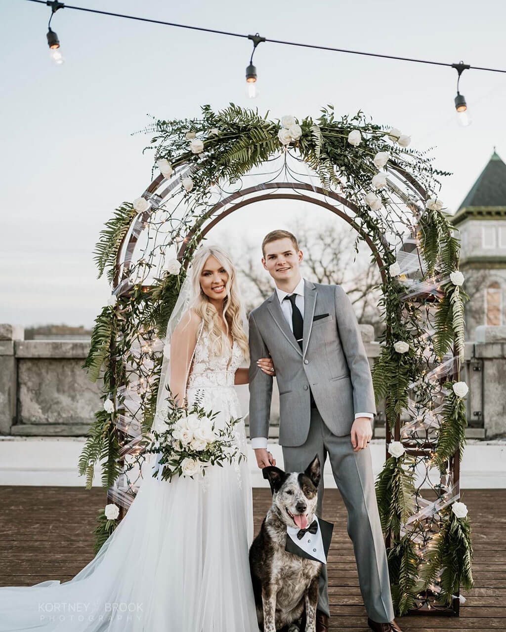 Bride and groom under a rose-and-fern arch on The Gaderian rooftop with their dog